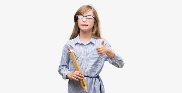 Young blonde child holding big pencil happy with big smile doing ok sign, thumb up with fingers, excellent sign - Photo, image