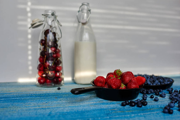 On a blue vintage wooden desk are two cast iron pans that sprinkling strawberries and blueberries near the scene along there are two glass bottles with milk and cherries - Foto, imagen