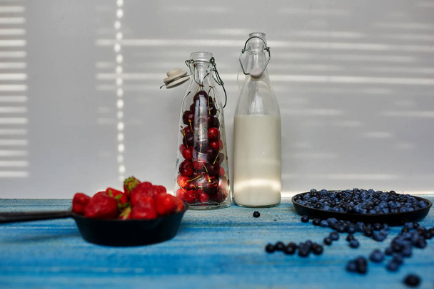 On the kitchen table are two bottles of a transparent glass with milk and the second with strawberries cherries next two pans in kototryh large sprinkling fresh berries - Foto, Imagem
