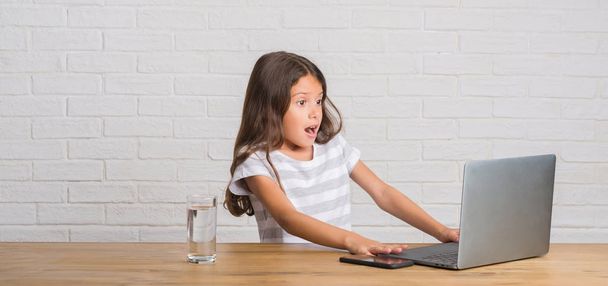 Молодой латиноамериканец, сидящий на столе с компьютером, напуганный в шоке с неожиданным лицом, напуганный и взволнованный выражением страха
 - Фото, изображение