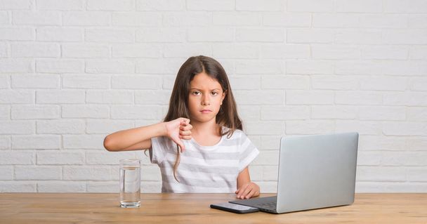 Jeune enfant hispanique assis sur la table à l'aide d'un ordinateur portable avec le visage en colère, signe négatif montrant aversion avec les pouces vers le bas, concept de rejet
 - Photo, image