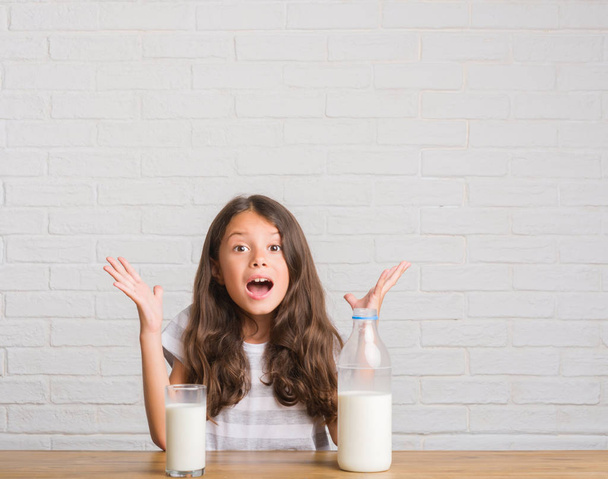 Nuori latino lapsi istuu pöydällä juoden lasillisen maitoa erittäin onnellinen ja innoissaan, voittaja ilme juhlii voittoa huutaen iso hymy ja nosti kädet.
 - Valokuva, kuva