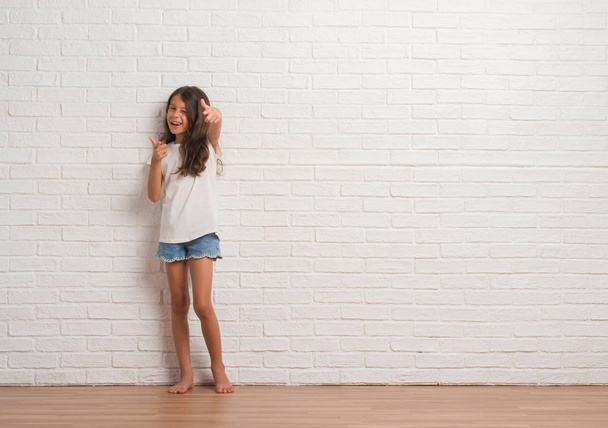 Joven niño hispano pisando la pared de ladrillo blanco señalando los dedos a la cámara con la cara feliz y divertida. Buena energía y vibraciones
. - Foto, Imagen