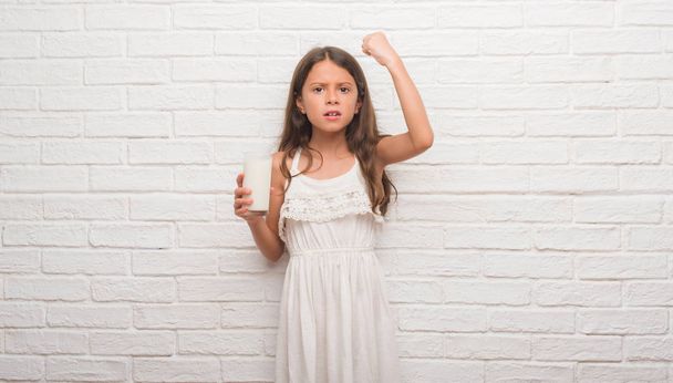 junges hispanisches Kind über weißer Ziegelwand, das ein Glas Milch trinkt, genervt und frustriert schreit vor Wut, verrückt und schreit mit erhobener Hand, Wut-Konzept - Foto, Bild