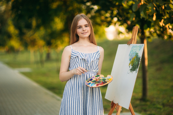 Hermosa artista chica está de pie en el parque delante de la imagen y la celebración de la paleta con pinturas
 - Foto, Imagen