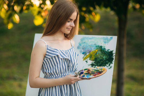 Beautiful girl artist is standing in the park and holding palette with paints - Photo, Image