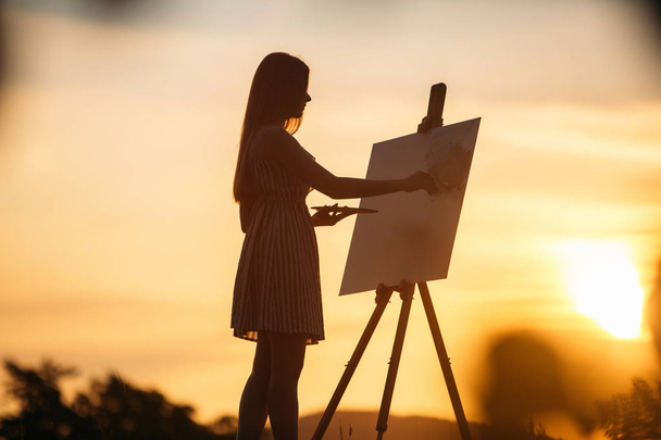 Silhouette einer blonden Künstlerin. Lady malt mit Hilfe von Farben ein Bild auf die Leinwand. Eine hölzerne Staffelei behält das Bild. Sommer ist ein sonniger Tag, Sonnenuntergang - Foto, Bild