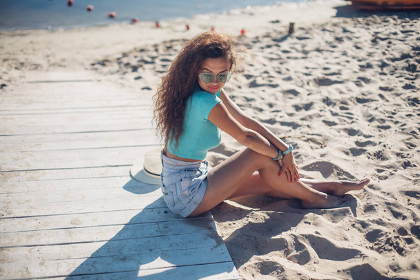 όμορφα σγουρά κορίτσι που κάθεται στην παραλία. παραθεριστικές κατοικίες διάθεση προβολή πλευρά πορτρέτο της μια χαλαρή γυναίκα σκέφτεται και να ψάχνει μακριά. - Φωτογραφία, εικόνα