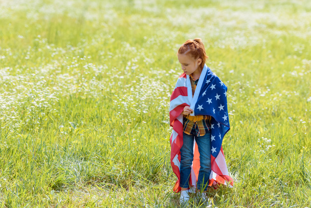 niño de pelo rojo cubierto de bandera americana de pie en el campo de verano
 - Foto, imagen