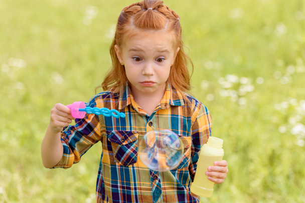 Porträt eines geschockten Kindes, das Seifenblasen auf einer Wiese pustet - Foto, Bild