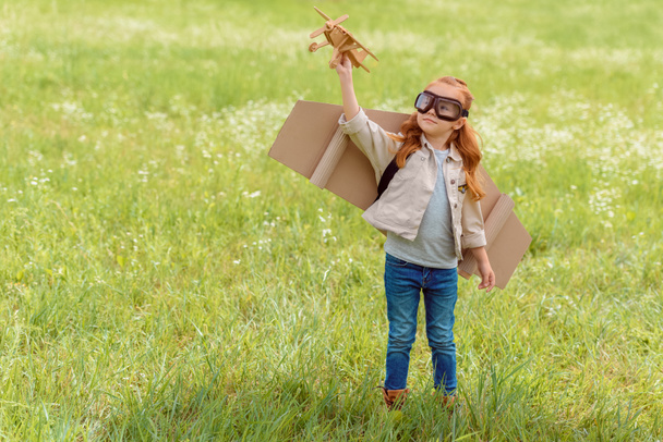 portrait of little child in pilot costume with wooden toy plane standing in meadow - Photo, Image