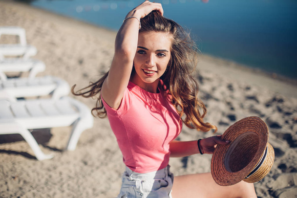 stijlvolle emotionele meisje draagt stro hoed poseren naast exotische zomer strand. Amazing brunette krullend vrouw zit op chaise-longue. Haren wapperen in de wind - Foto, afbeelding