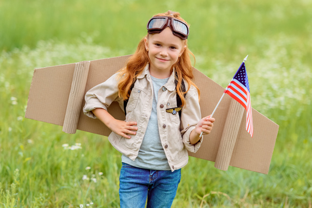 portrait of smiling kid in pilot costume with american flagpole standing in meadow - Photo, Image