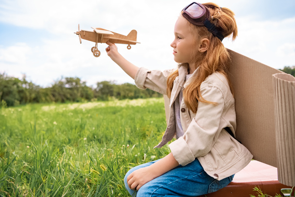 bambino in costume da pilota con aereo giocattolo in legno in mano seduto su valigia retrò in campo
 - Foto, immagini