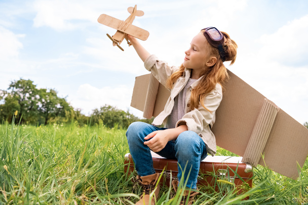 kid in pilot costume with wooden toy plane in hand sitting on retro suitcase in field - Photo, Image