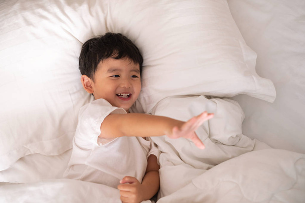 3 years old little cute Asian boy in white shirt at home on the bed, kid lying playing and smiling on white bed with pillow and blanket, top view with copy space for add text. - Foto, Imagen