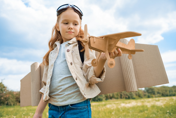 retrato de niño de pelo rojo en traje piloto sosteniendo avión de madera en el campo de verano
 - Foto, Imagen