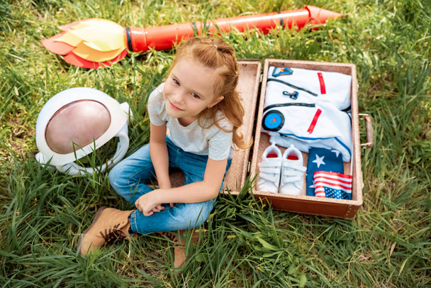 high angle view of smiling red hair kid sitting near suitcase with cosmonaut costume and rocket on green grass - Fotó, kép