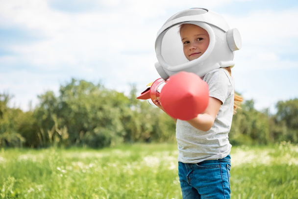 Porträt eines Kindes mit Kosmonautenhelm und Rakete in den Händen im Feld stehend - Foto, Bild