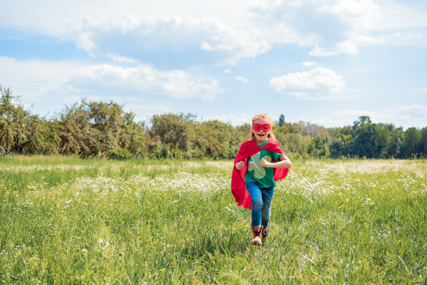 happy kid in red superhero cape and mask running in meadow on summer day - Photo, Image