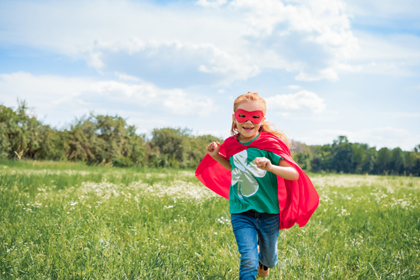 happy kid in red superhero cape and mask running in meadow on summer day - Photo, Image