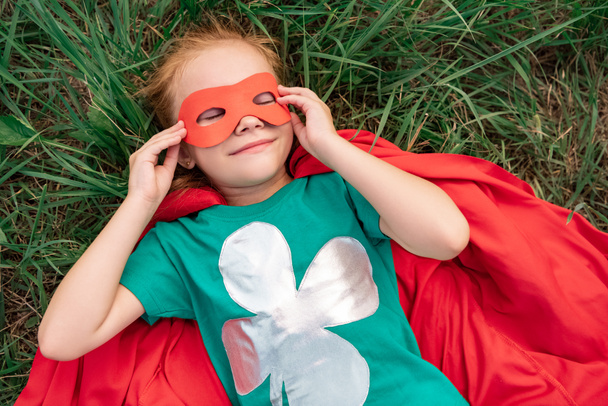 bovenaanzicht van het kind met de ogen gesloten in rode superheld cape en masker liggend op groen gras - Foto, afbeelding