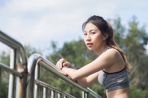Young woman relaxing after jogging exercise on fence at park to freshen her body and enjoy warm light in morning. Young asian woman take a break from running execise. Outdoor exercise activity. - Photo, image