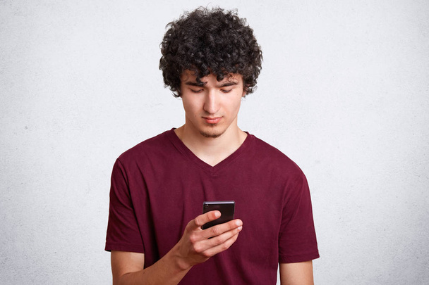 Good looking guy with concetrated look in mobile phone, recieves message with offer to go out, wears casual t shirt, poses against white concrete wall. Curly man uses modern technologies indoor - Foto, Imagem
