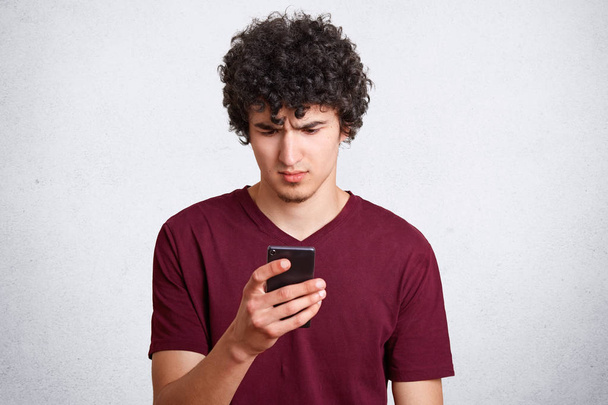 Portrait of attractive curly male looks in bewilderment on mobile phone, reads news via cellular, dressed casually, poses against white concrete wall. People, youth, technology and lifestyle concept - Photo, image