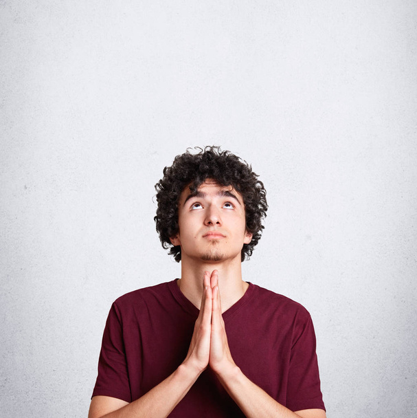 vertikale Aufnahme eines angenehm aussehenden elenden Mannes betet um etwas, blickt hoffnungsvoll nach oben, bittet um Gesundheit, posiert vor weißem Hintergrund mit Leerraum für Ihre Werbeinhalte - Foto, Bild
