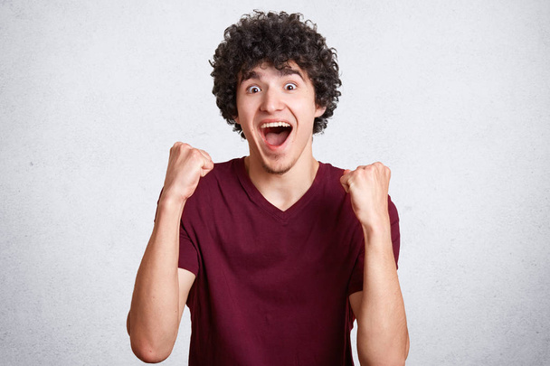 Emotive overjoyed male teenager clenches fists, has curly dark hair, wears casual t shirt, poses against white background. Headshot of glad successful man succeed in something. Wow, I did it! - Fotografie, Obrázek