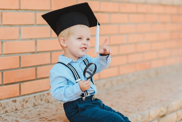 Little boy in spectacles and academic hat standing over against a brick wall of a school. Holds a magnifying glass and studies the environment - Foto, Imagem
