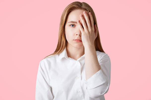 Attractive female covers face with hand, has tired expression, being fatigue and bored of daily routine, wears white shirt, isolated on pink background. People, facial expressions and emotions concept - Foto, immagini