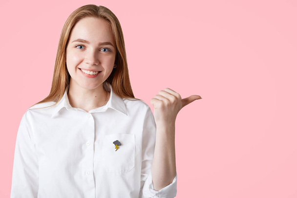 Horizontal portrait of happy Caucasian female indicates with thumb at blank copy space against pink background for your advetisement or promotional text, has positive expression, warm smile. - Foto, afbeelding