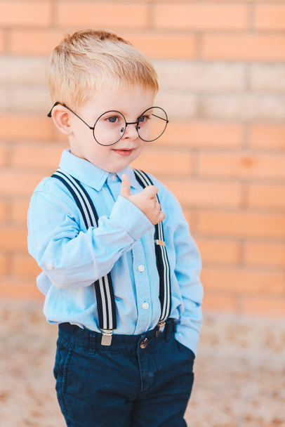 Back to school. Happy little boy in glasses with backpack and books against brick wall of the school building. - 写真・画像