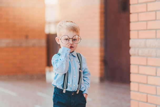 Powrót do szkoły. Szczęśliwy mały chłopiec w okularach z plecaka i książek przed murem budynku szkoły. - Zdjęcie, obraz