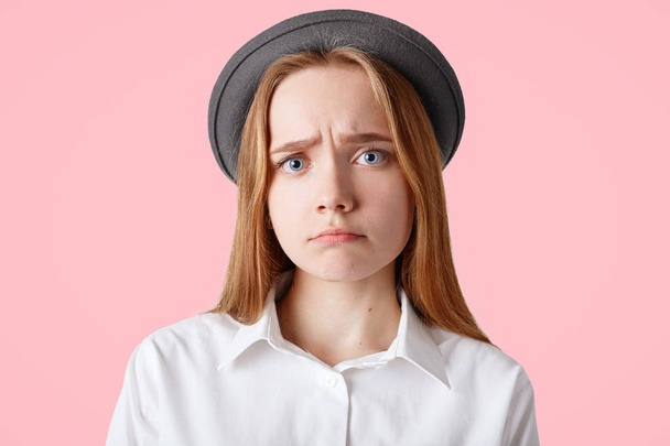 Studio shot of pleasant looking female with upset look, wears elegant hat, curves lips, has sorrowful expression, poses against pink background. Beautiful woman feels depressed after failure - Photo, Image