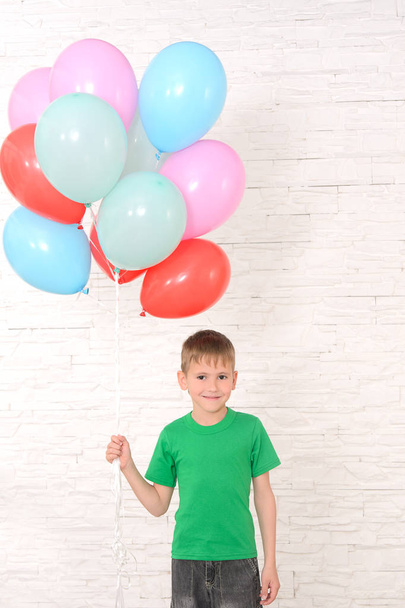 boy in green t-shirt holding a lot of colorful balloons against a white brick wall - Фото, изображение