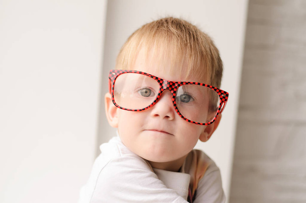 blonder Junge mit roter Brille in Großaufnahme, sitzend auf einem Hintergrund aus weißem Backstein - Foto, Bild