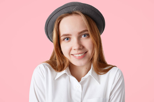 Headshot of pleased female has healthy skin, pleasant smile on face, wears elegant black hat, isolated on pink studio background. Lovely woman expresses positive emotions, models indoor alone - Foto, afbeelding
