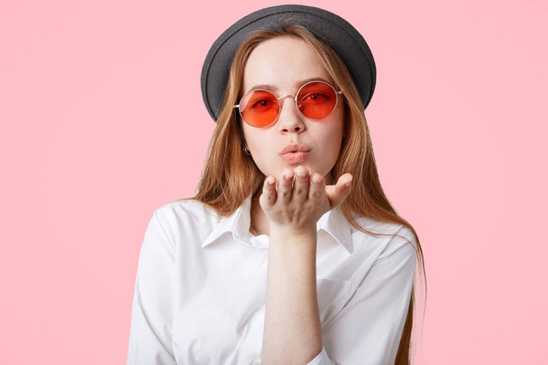Lovely fashionable hipster girl wears trendy pink shades and black hat, blows air kiss at camera, poses against pink background, expresses her love to surrounding, Pleasant looking female flirts - Foto, Imagen