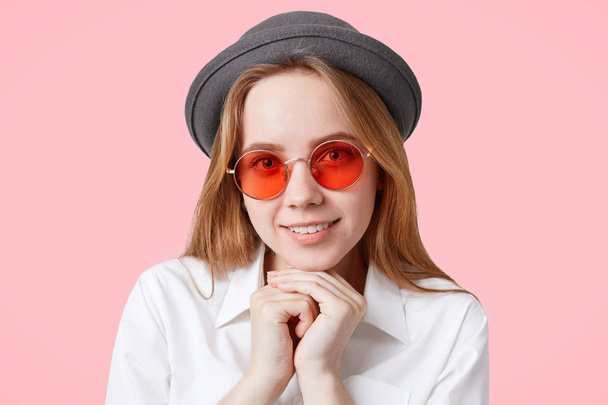 Portrait of happy delighted female model in round red sunglasses and elegant hat, keeps hands under chin, smiles broadly, isolated over pink background. People, style and facial expressions. - Fotoğraf, Görsel