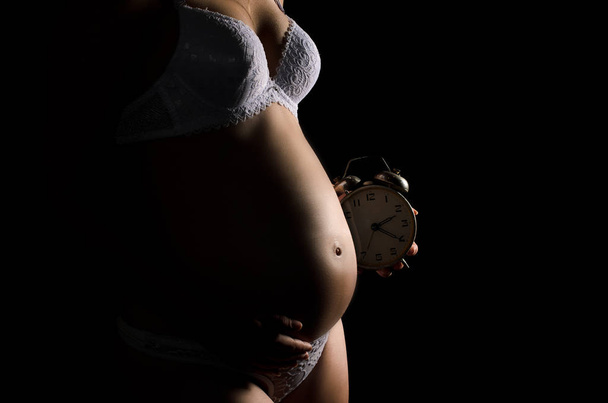Closeup nahé ženy těhotné břicho, jednou rukou drží jeho žaludek, dugaya - hodin na zcela černém pozadí - Fotografie, Obrázek