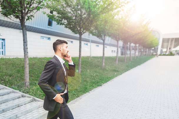 Young smiling businessman talking on cell phone and walking near modern office building, copy space. Business, technology and communication concept - Foto, Imagen