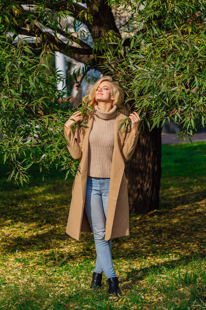 Beautiful elegant blonde woman dressed in a coat standing under the tree in autumn park. Sunny october day. - Foto, afbeelding