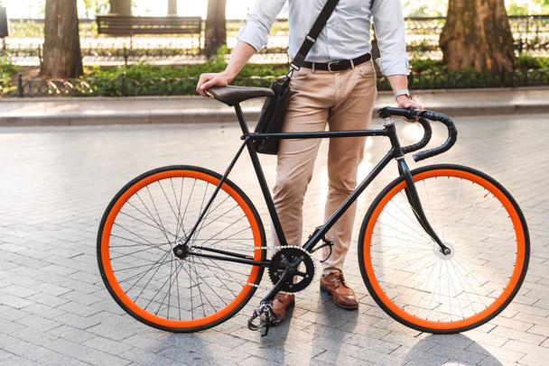 Cropped image of a young man in shirt with bag standing with a bicycle on a city street - Foto, Imagen