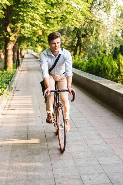 Portrait of a smiling young man dressed in shirt with bag riding on a bicycle outdoors - Foto, imagen