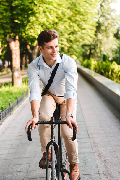 Portrait of a happy young man dressed in shirt with bag riding on a bicycle outdoors - Foto, imagen