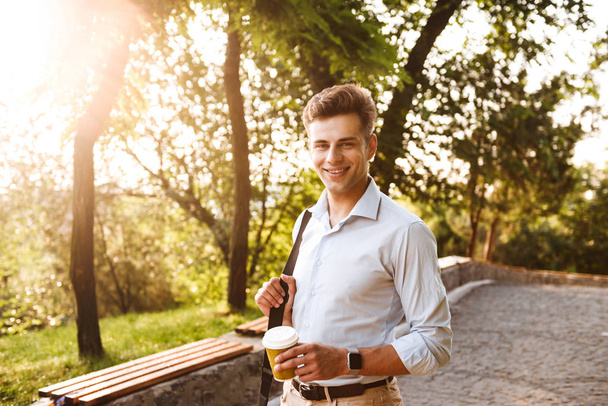 Portrait of a smiling young man dressed in shirt holding cup of coffee while standing outdoors - Fotoğraf, Görsel