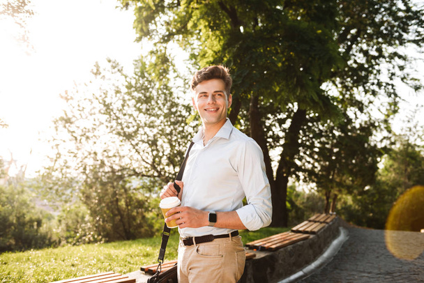 Happy young man in shirt walking at the city park with cup of coffee - Photo, image